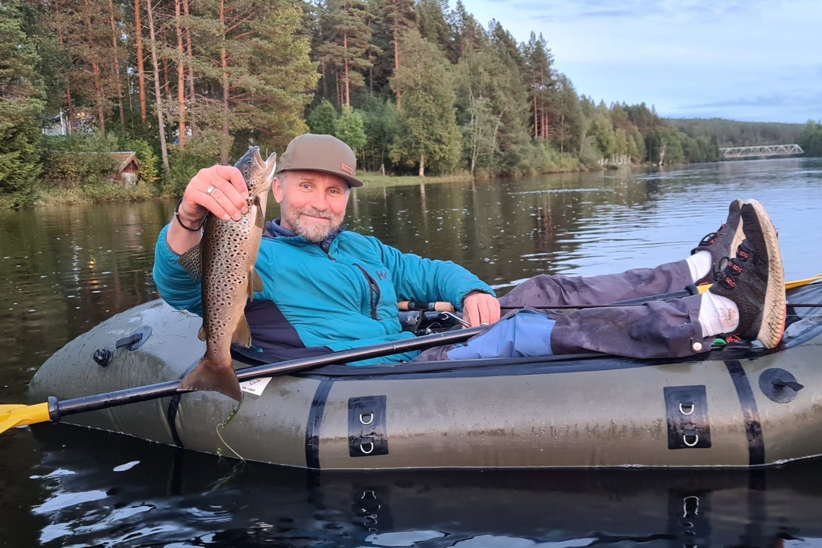 An angler relaxing in a packraft on a calm river, holding a brown trout