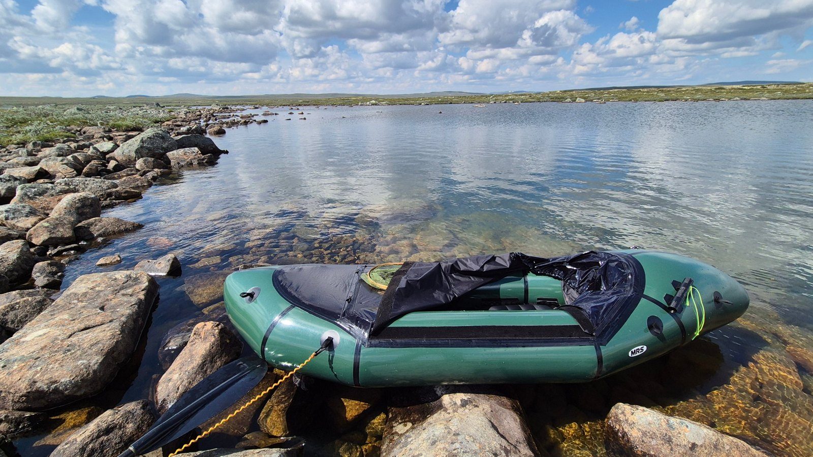 A green packraft and paddle resting on rocks by clear water